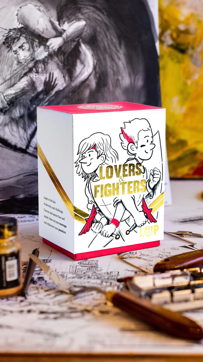 Living & Powerful 'Lovers & Fighters' boxed card set, gold-foiled, on a desk with hand-drawn artwork behind it